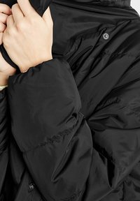 Svart puffjacka med en slät, glänsande finish. Har hög krage, fast huva och knappstängningar. Tjockt, isolerat tyg med quiltat mönster.