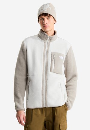 Homme portant une veste polaire beige et blanche avec une poche poitrine zippée et un bonnet tricoté beige, pantalon cargo marron, debout, la main dans la poche.