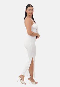 Vestido blanco ajustado con una abertura, tirantes finos ajustables y un detalle de nudo en la parte delantera. Combinado con sandalias de tacón alto en beige.