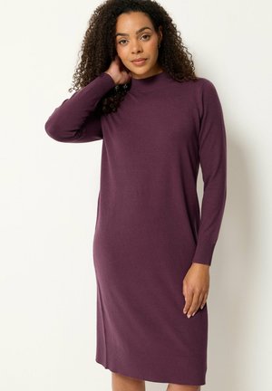 Femme aux cheveux bouclés portant une robe pull violette à manches longues, longueur genou, debout avec une main touchant son cou.