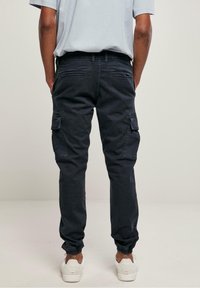 Homme portant un pantalon cargo noir avec des poignets élastiques et des baskets blanches, debout, dos tourné, sur un fond uni.