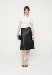 Black leather A-line skirt with pleats, paired with a fitted white short-sleeve top. Model holds a small black handbag, wearing strappy heels.