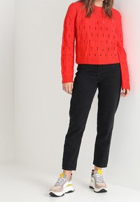 Červený pletený svetr s texturovaným vzorem, kombinovaný s černými džíny s rovnými nohavicemi a vícebarevnými robustními teniskami s žlutými akcenty.