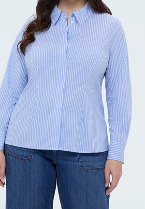 Mujer que lleva una camisa de botones de manga larga, de color azul claro con rayas verticales, combinada con jeans vaqueros azul oscuro.