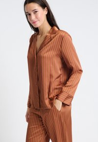 Brązowy satynowy zestaw piżamowy z różowymi pionowymi paskami, składający się z koszuli na guziki oraz pasujących spodni. Koszula ma wycięty kołnierzyk i kieszenie.