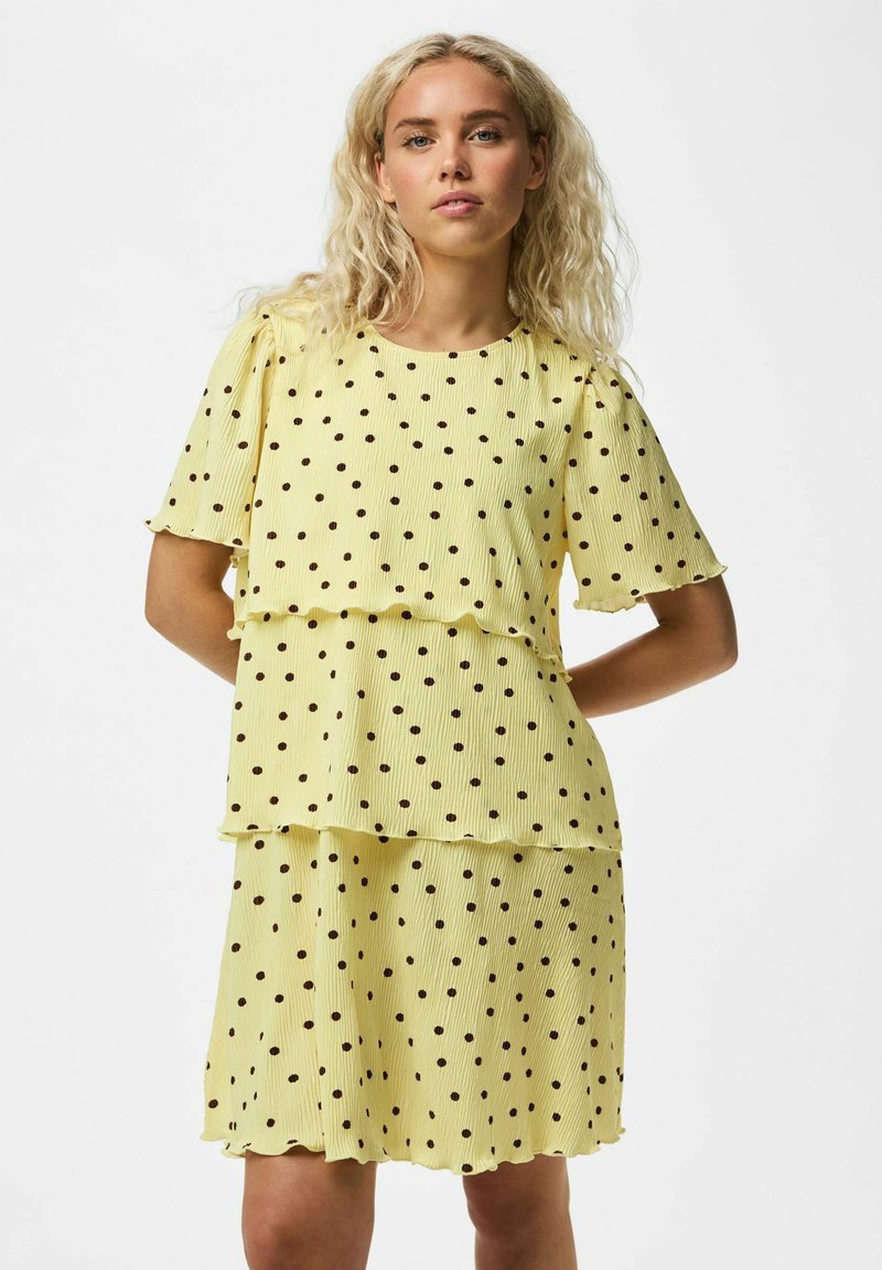 Jeune femme aux cheveux blonds ondulés portant une robe jaune à manches courtes avec des pois noirs, debout sur un fond blanc uni.