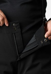Handen die de tailleband en het rits van een zwarte broek openhouden, waarbij metalen drukknoopjes en ritsdetails zichtbaar zijn.