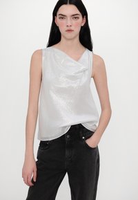 Haut sans manches en argent brillant avec un design drapé, associé à un jean noir taille haute. Légent éclat sur le tissu lisse, créant un aspect texturé.