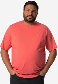 Koraliowy t-shirt z dekoltem w serek wykonany z miękkiego materiału, z krótkimi rękawami i subtelnie wytłoczonym logo na piersi, zestawiony z ciemnymi dżinsami.