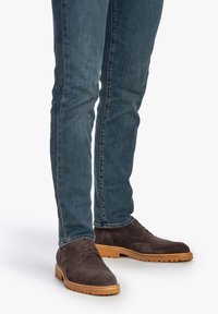 Zapatos de gamuza marrón con un diseño de cordones, que cuentan con una suela de goma y perforaciones sutiles, combinados con unos jeans azules ajustados.