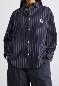 Camisa de botones azul marino con estrechas rayas verticales blancas, un bolsillo en el pecho y una etiqueta de tela en el bolsillo. Material de algodón suave.