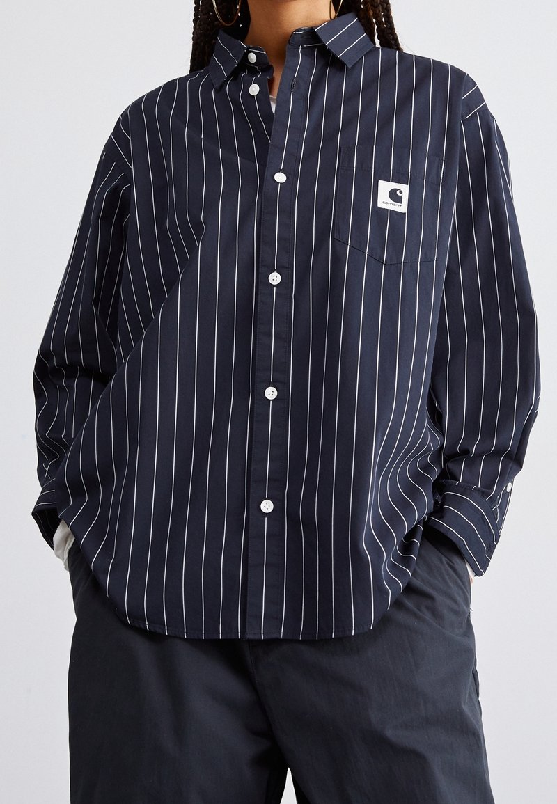 Camisa de botones azul marino con estrechas rayas verticales blancas, un bolsillo en el pecho y una etiqueta de tela en el bolsillo. Material de algodón suave.