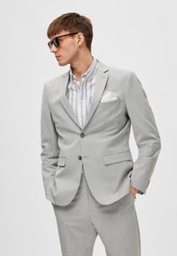 Traje gris claro de corte ajustado, con solapa de muesca y dos botones. Debajo, una camisa a rayas, complementada con un pañuelo de bolsillo blanco.