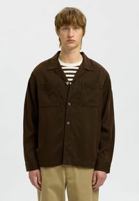 Brązowa, zapinana na guziki kurtka z lekkiego materiału, z dwoma kieszeniami na piersi, wykładanym kołnierzem i luźnym krojem, noszona na koszulę w paski.