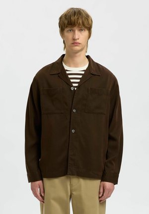 Pruun, nööpidega jakk kergest materjalist, millel on kaks rinna taskut, lõikatud krae ja mugav lõige, kantud triibulise särgi üle.