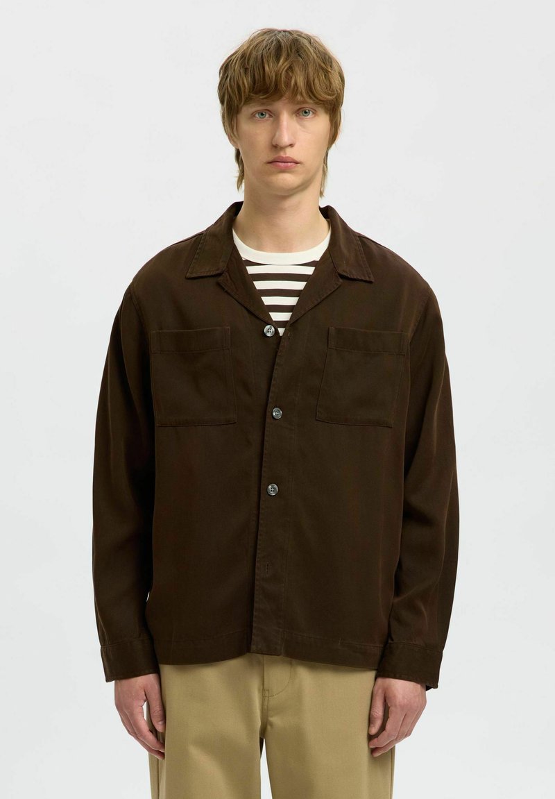 Brązowa, zapinana na guziki kurtka z lekkiego materiału, z dwoma kieszeniami na piersi, wykładanym kołnierzem i luźnym krojem, noszona na koszulę w paski.