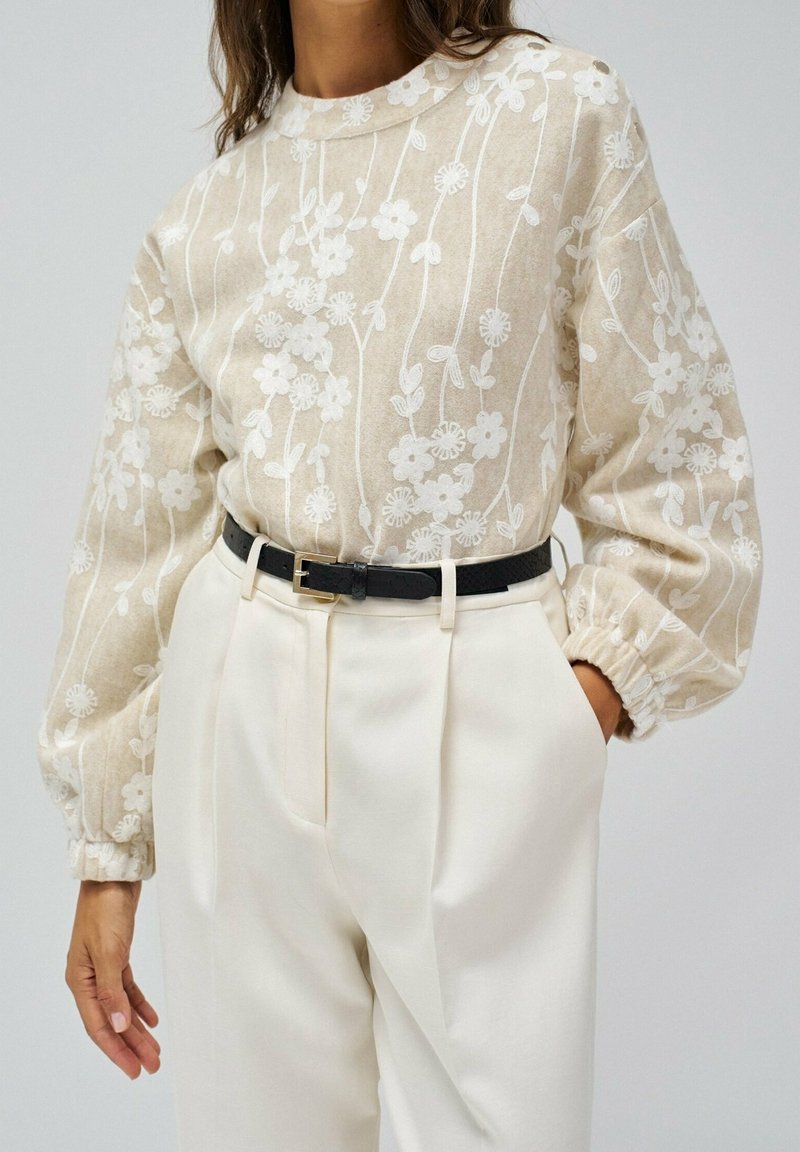 Pull beige avec broderie florale blanche rentré dans un pantalon blanc taille haute, ceinture noire, main gauche dans la poche, main droite détendue le long du corps.