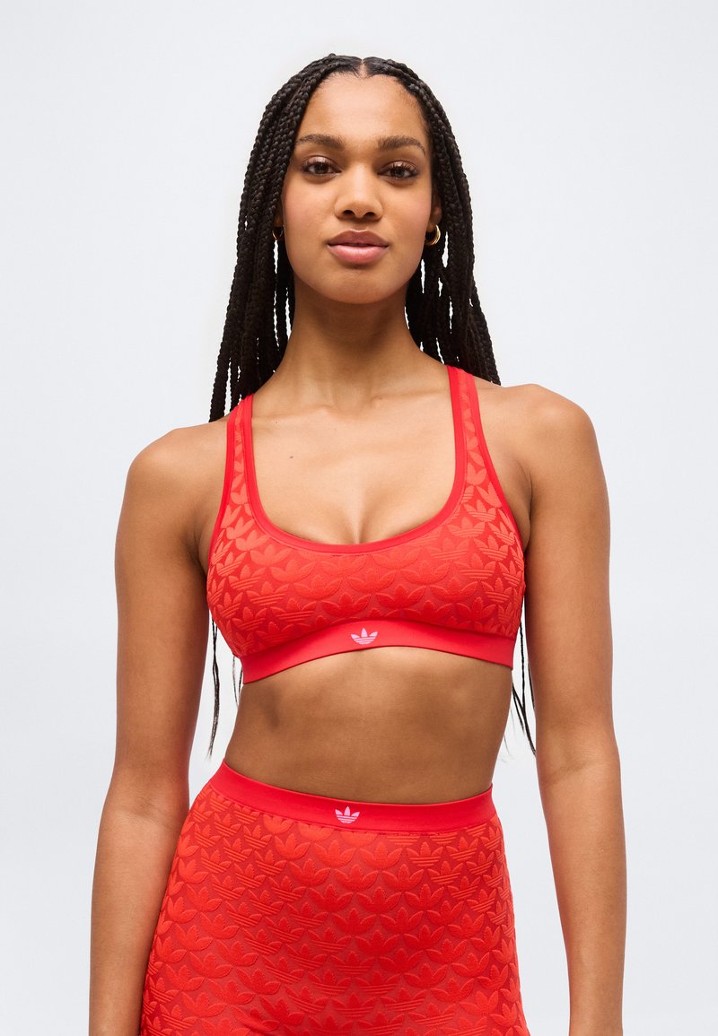 Νέα γυναίκα με μακριά κοτσίδα, φοράει αθλητικό σουτιέν με κόκκινο σχέδιο και αντίστοιχο κολάν ψηλής μέσης, κοιτάει προς τα εμπρός με απλό φόντο.