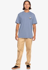 Light blue cotton t-shirt with short sleeves, front chest logo, paired with khaki pants and black sneakers. Smooth texture, relaxed fit.