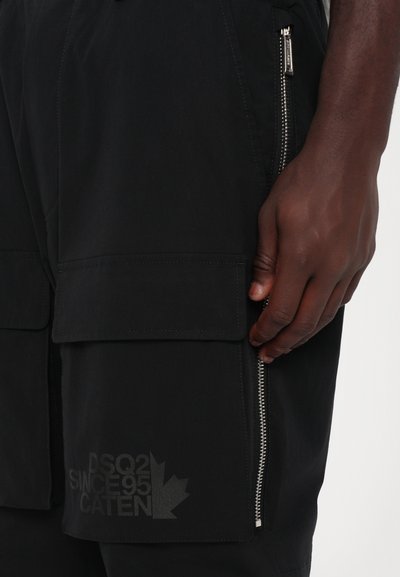 Black cargo shorts with zippered pockets, featuring a smooth texture and an embossed logo detail on the leg. Positioned hand-on-pocket view.