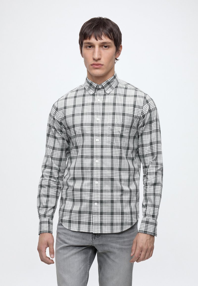 Ung mand med kort mørkt hår iført en grå og hvid ternet skjorte med knapper og lysegrå jeans mod en ensfarvet hvid baggrund.