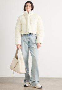 Zastrihnutá biela fleecová bunda s vysokým golierom, kombinovaná so svetlomodrými rozšírenými džínsami a béžovou taškou. Tenisky dopĺňajú outfit.