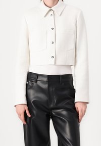 Veste blanche courte avec un tissage texturé, deux poches avant, boutons argentés et col pointu, associée à un pantalon en cuir noir.