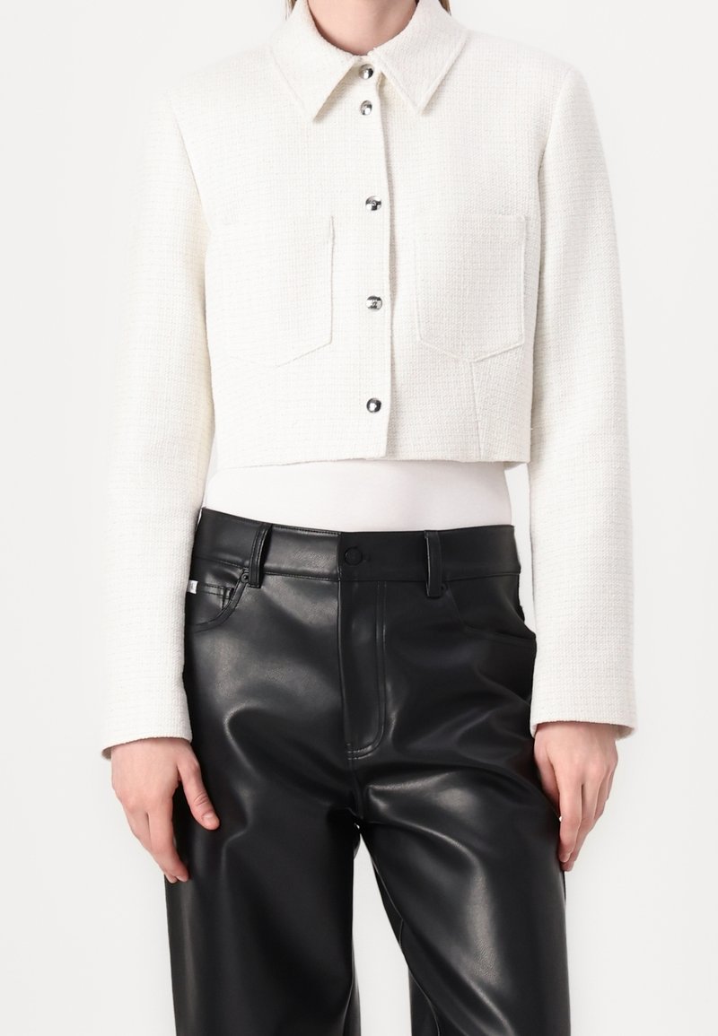 Veste blanche courte avec un tissage texturé, deux poches avant, boutons argentés et col pointu, associée à un pantalon en cuir noir.