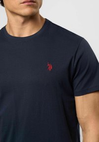 Férfi sötétkék környakú pólót visel, amelyen a bal mellrészen egy kis piros hímzett logó látható, amely egy lándzsát fogó alakot ábrázol.