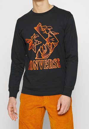 Osoba nosząca czarną koszulkę z długim rękawem z pomarańczową grafiką góry i drzew oraz tekstem "CONVERSE", zestawioną z jaskrawo pomarańczowymi sztruksowymi spodniami.
