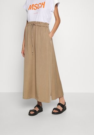 Persona con una falda larga beige de cintura alta y cinturón atado, camiseta blanca de MSCH y sandalias negras abiertas, de pie con una mano en el bolsillo.