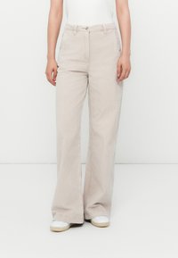 Persona che indossa pantaloni in corda beige a gamba ampia, sneakers bianche e una maglietta bianca infilata, in piedi contro uno sfondo bianco uniforme.