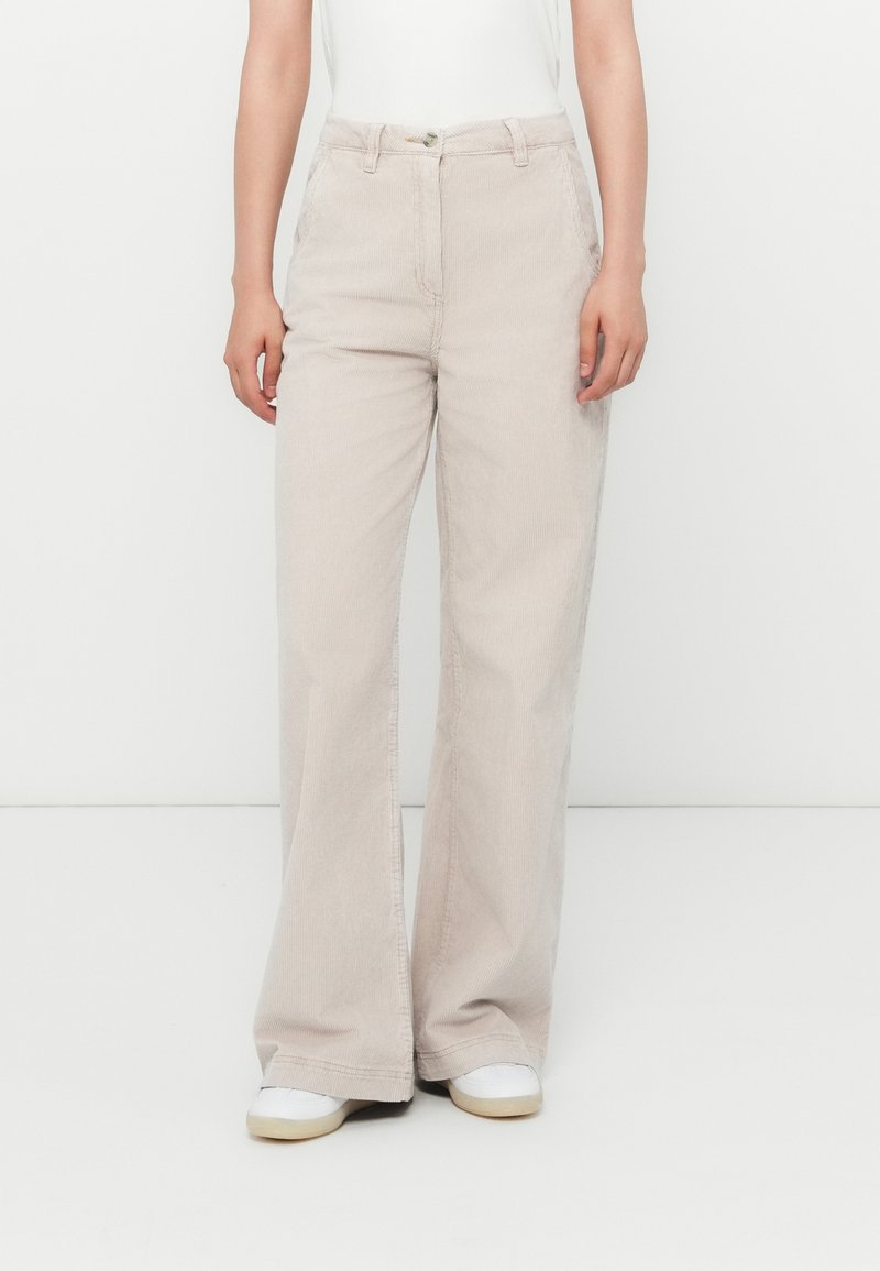 Persona che indossa pantaloni in corda beige a gamba ampia, sneakers bianche e una maglietta bianca infilata, in piedi contro uno sfondo bianco uniforme.