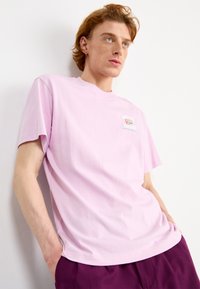 T-shirt di cotone rosa chiaro con collo rotondo, maniche corte e un piccolo logo sul petto. Il modello indossa pantaloni viola, con le mani in tasca.