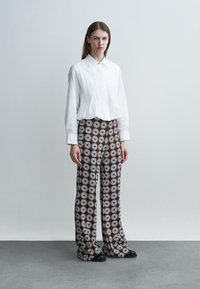 Moteris stovi vilkėdama laisvą baltą ilgomis rankovėmis marškinių ir platėjantų kelnių rudos spalvos fone su baltų ramunėlių raštu.