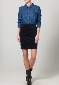 Camicia di jeans con bottoni e due tasche sul petto, abbinata a una gonna a tubo in velluto a costine blu scuro. Stivaletti neri completano il look.