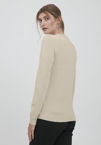 Beige neule, jossa on hunajakenno-kuvio, resorit hihoissa ja helmaraita. Mallia on kuvattu selkäpuolelta, neutraalilla taustalla.