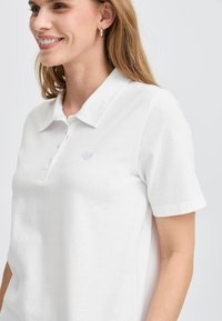 Biely polo s krátkymi rukávmi, golierom a jemným vyšívaním srdca na hrudi. Mäkký bavlnený materiál s čistou, hladkou textúrou.