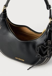 Μαύρη δερμάτινη τσάντα χειρός με λευκά ραφτά, χρυσό λογότυπο "LOVE MOSCHINO" και ένα καρδιόσχημο γούρι, με εσωτερική θήκη με φερμουάρ.