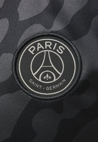 Musta kangas, jossa on hienovarainen maastokuvio, ja siinä on ympyrän muotoinen, kohokuvioitu merkki, jossa lukee "PARIS SAINT-GERMAIN" ja joka sisältää liljan symbolin.