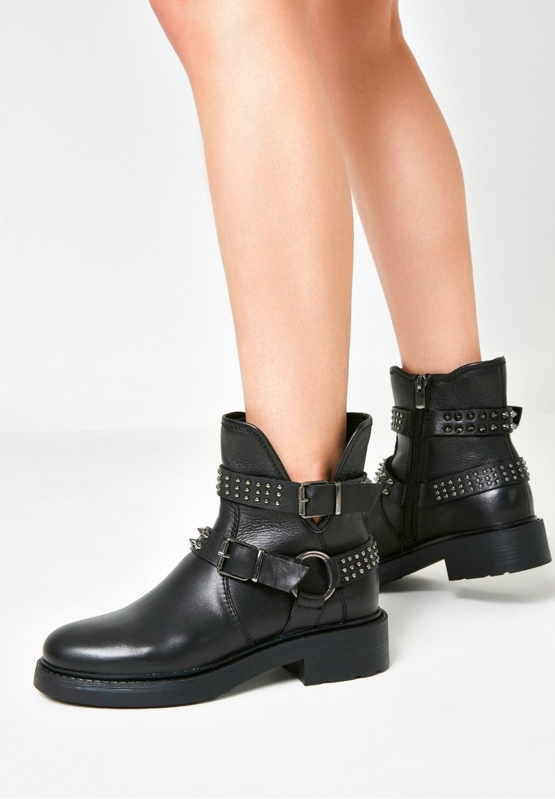 Bottes courtes en cuir noir ornées de clous argentés, de boucles et de fermetures éclair latérales, portées sur des jambes nues sur un fond blanc uni.