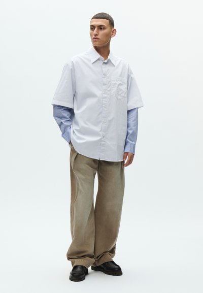 Mladý muž stojí, oblečený v modrej pruhovanej vrstvenej košeli s dlhým rukávom pod bielou krátkym rukávom s gombíkmi, voľné hnedé nohavice a čierne topánky.