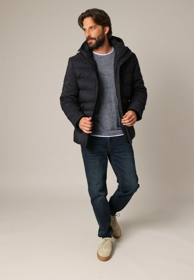 Homme barbu ajustant une veste en doudoune noire, portant un pull gris, un jean bleu foncé et des chaussures à lacets beiges, se tenant sur un fond neutre.