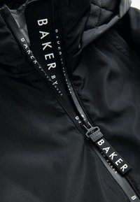 Černá bunda na zip s hladkým, lesklým vnějším povrchem. Výrazné logo na jezdcích zipu a prošívaný detail na vnitřní straně.