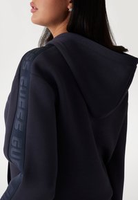 Sweat à capuche marine avec une coupe décontractée, doté d'une capuche avec cordon de serrage et d'une bande logo texturée le long de la manche. Tissu lisse, design minimaliste.