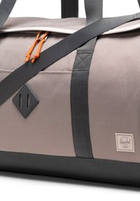 Duffelbag aus taupefarbenem Canvas mit schwarzem Boden, orangefarbenen Kordeln und schwarzen Trägern. Verfügt über einen Diamant-Patch und strukturierte Akzente.
