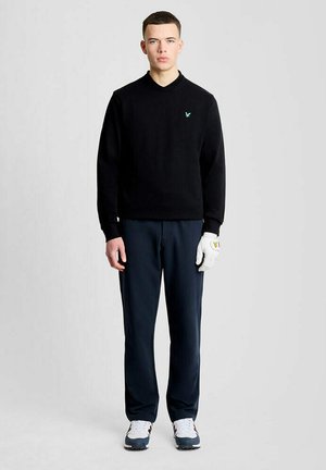 Ung mand iført sort sweater, mørke bukser, hvid golfhandske på venstre hånd og hvide sneakers står foran en ensfarvet lys baggrund.