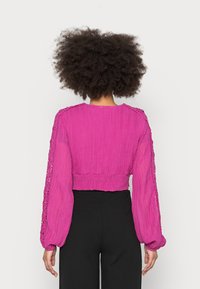 Fuchsia cropped blouse met lange, wijd uitlopende mouwen, gestructureerde stof en kantdetails langs de mouwen en de halslijn. Zwarte broek zichtbaar.