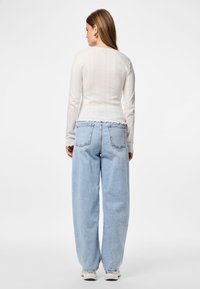 Mujer de espaldas vistiendo una blusa blanca de manga larga con canalé, unos vaqueros holgados de color azul claro, zapatillas blancas y pendientes de aro plateados sobre un fondo blanco.