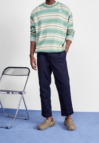 Zelená a proužkovaná mikina v pastelových tónech, námořnické kalhoty a béžové dřeváky. Má pohodlný střih a texturovanou látku. Vedle je chromová židle.